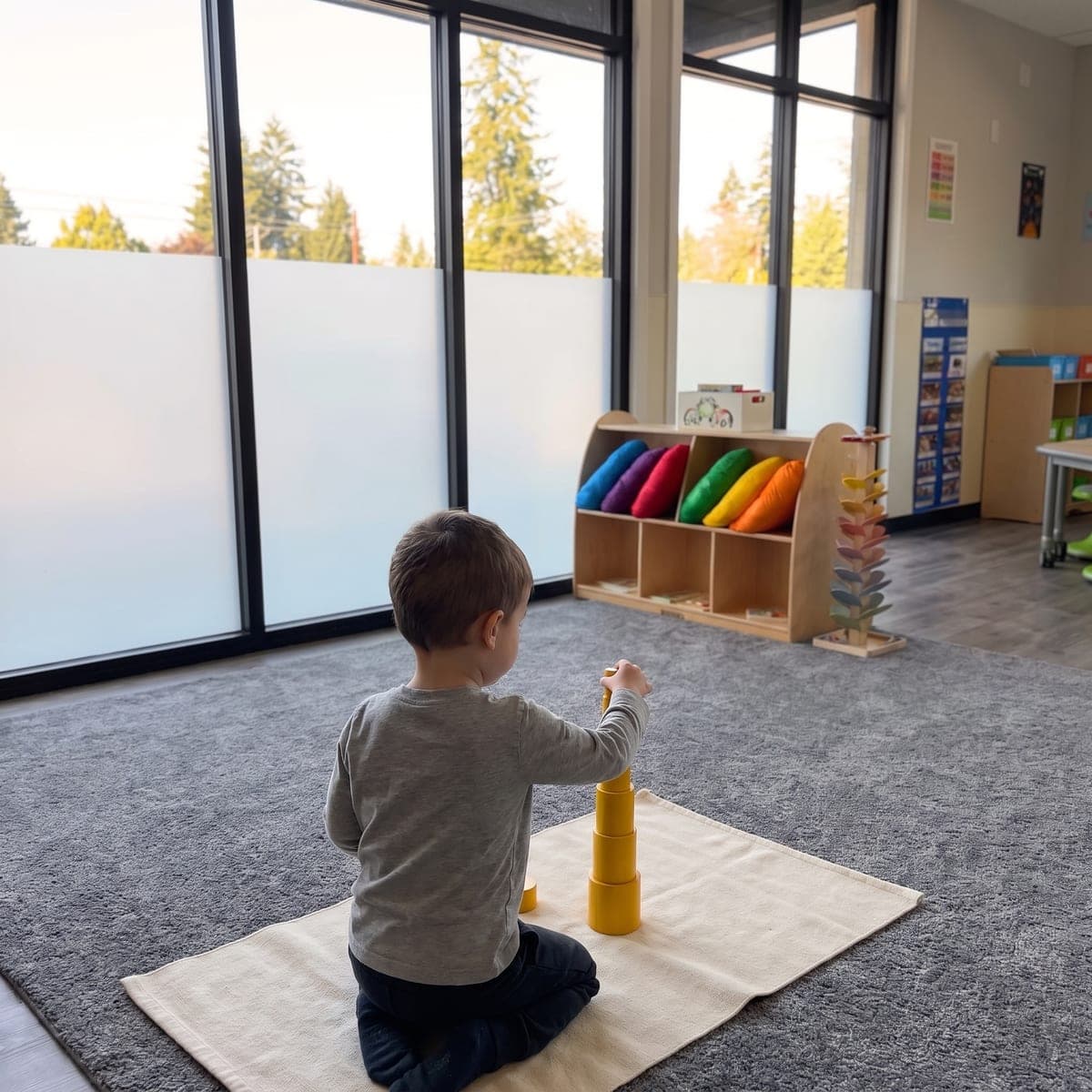 A child, seen from behind, stacking knobless cylinders into a tower on a work mat by the window