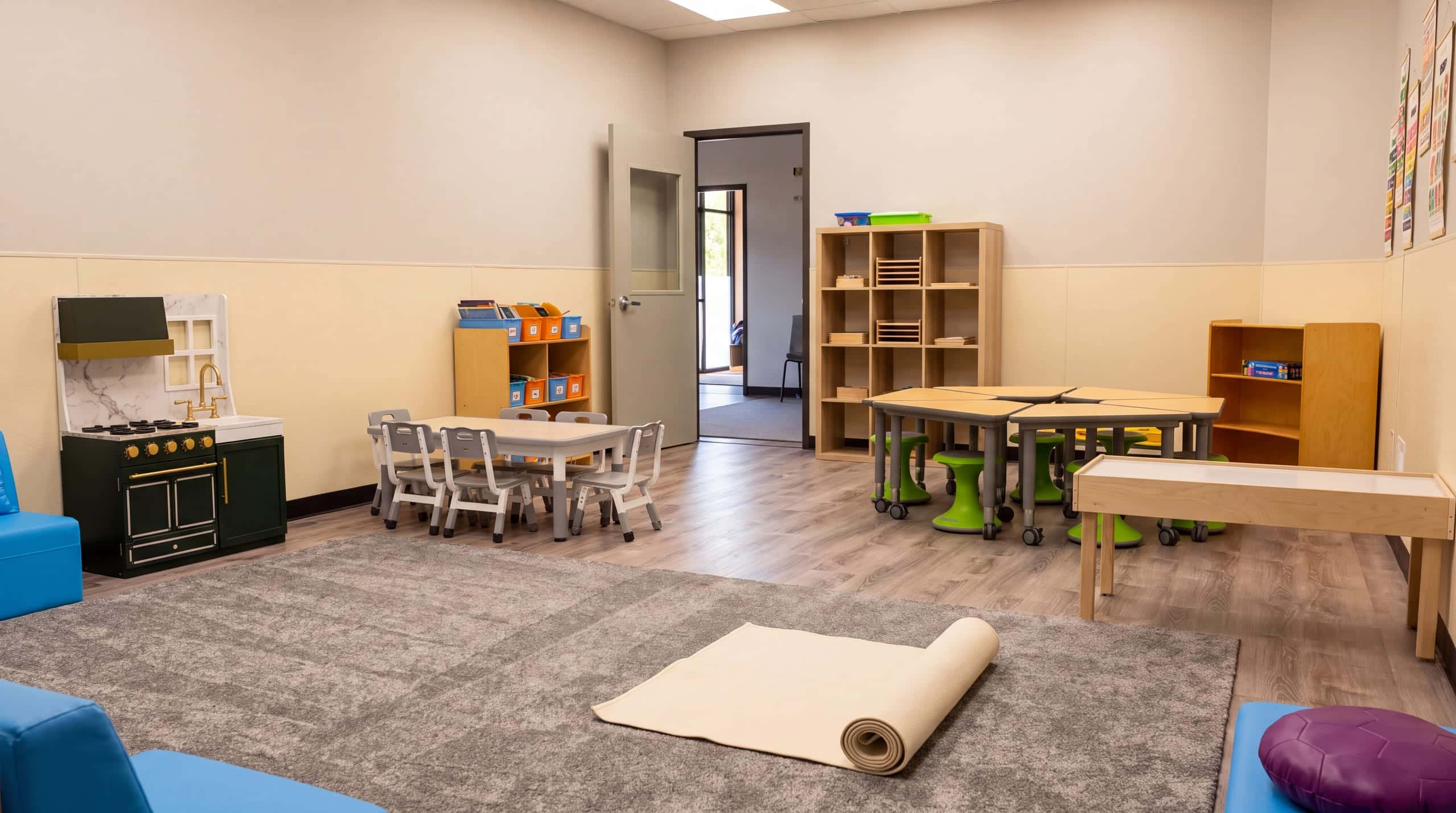 A classroom seen from a child's eye height: the soft grey rug, small tables, the play kitchen and low shelves