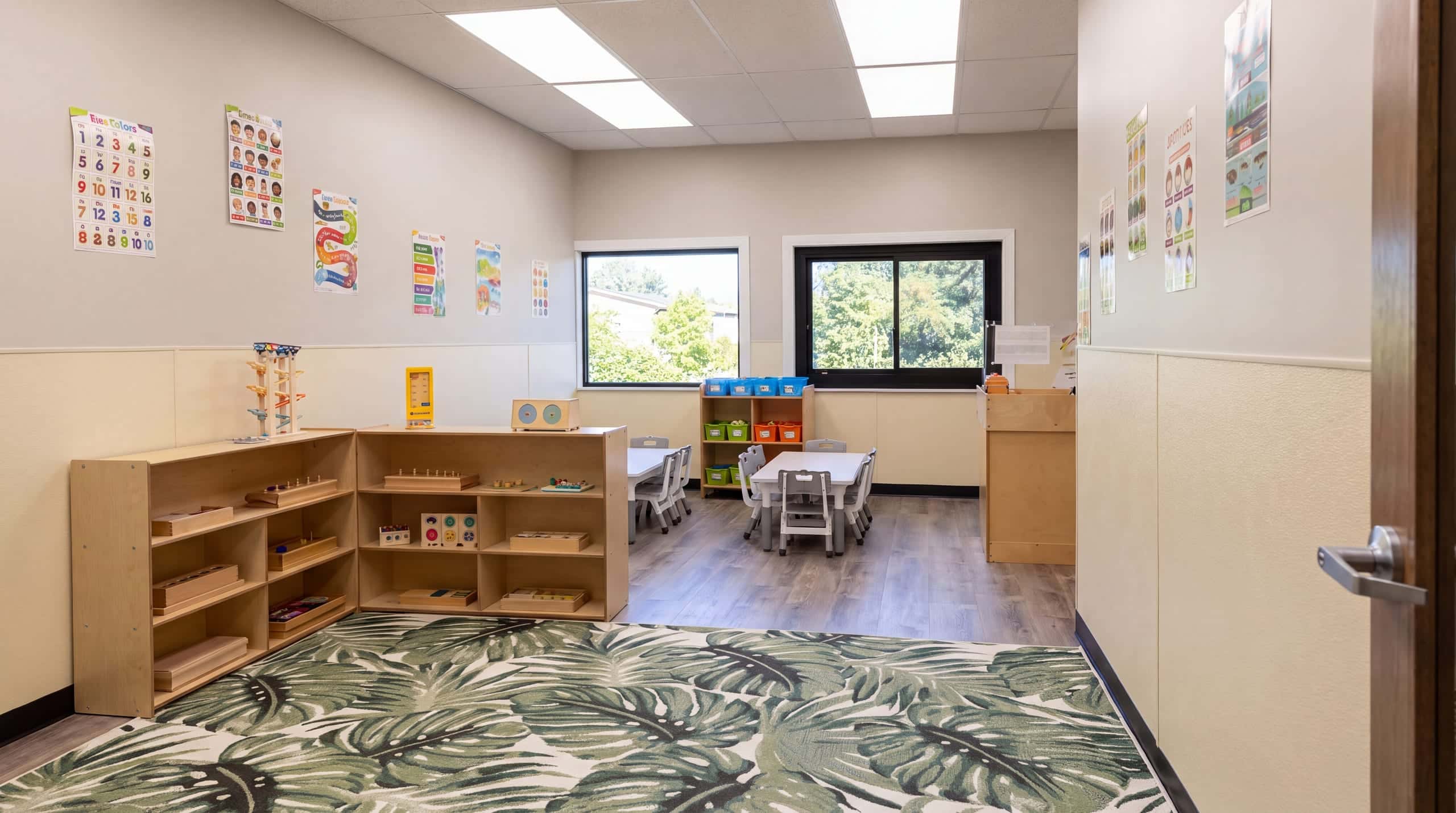 A classroom looking toward the windows: birch shelves of wooden materials, small tables, trees beyond the glass