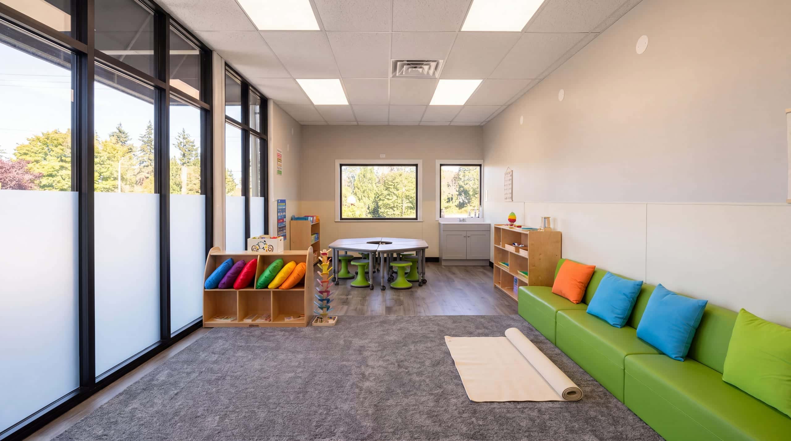 A classroom in soft daylight: tall windows along one wall, the green sofa, birch shelves and the charcoal rug, empty before the day begins