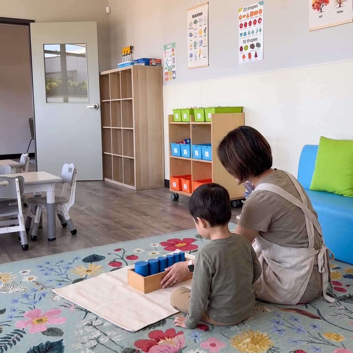 A guide and a child, seen from behind, side by side on a work mat during a one-to-one lesson