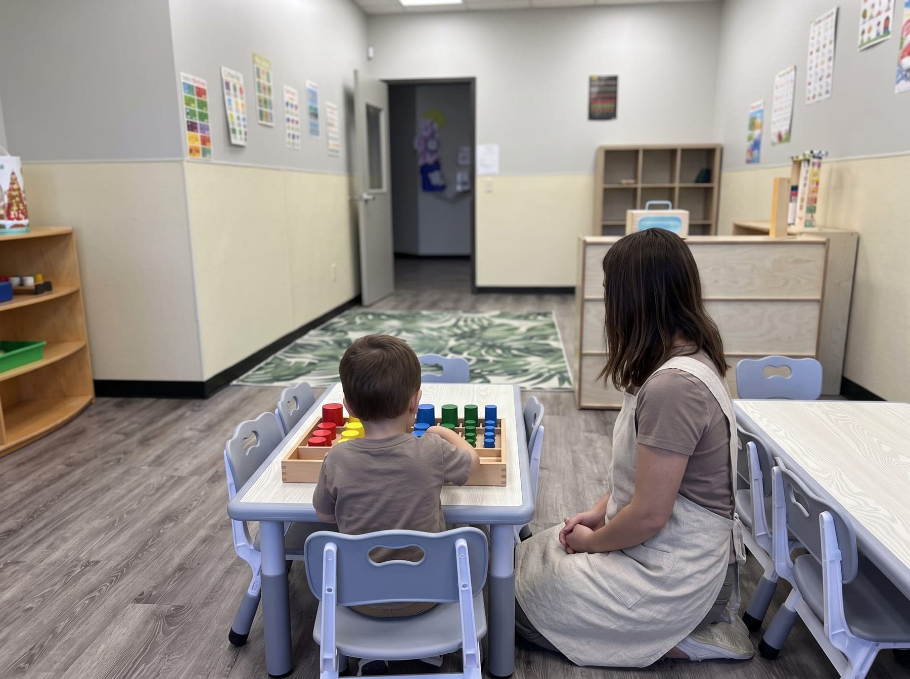 A guide, seen from behind, kneeling beside a child at a low table and quietly observing