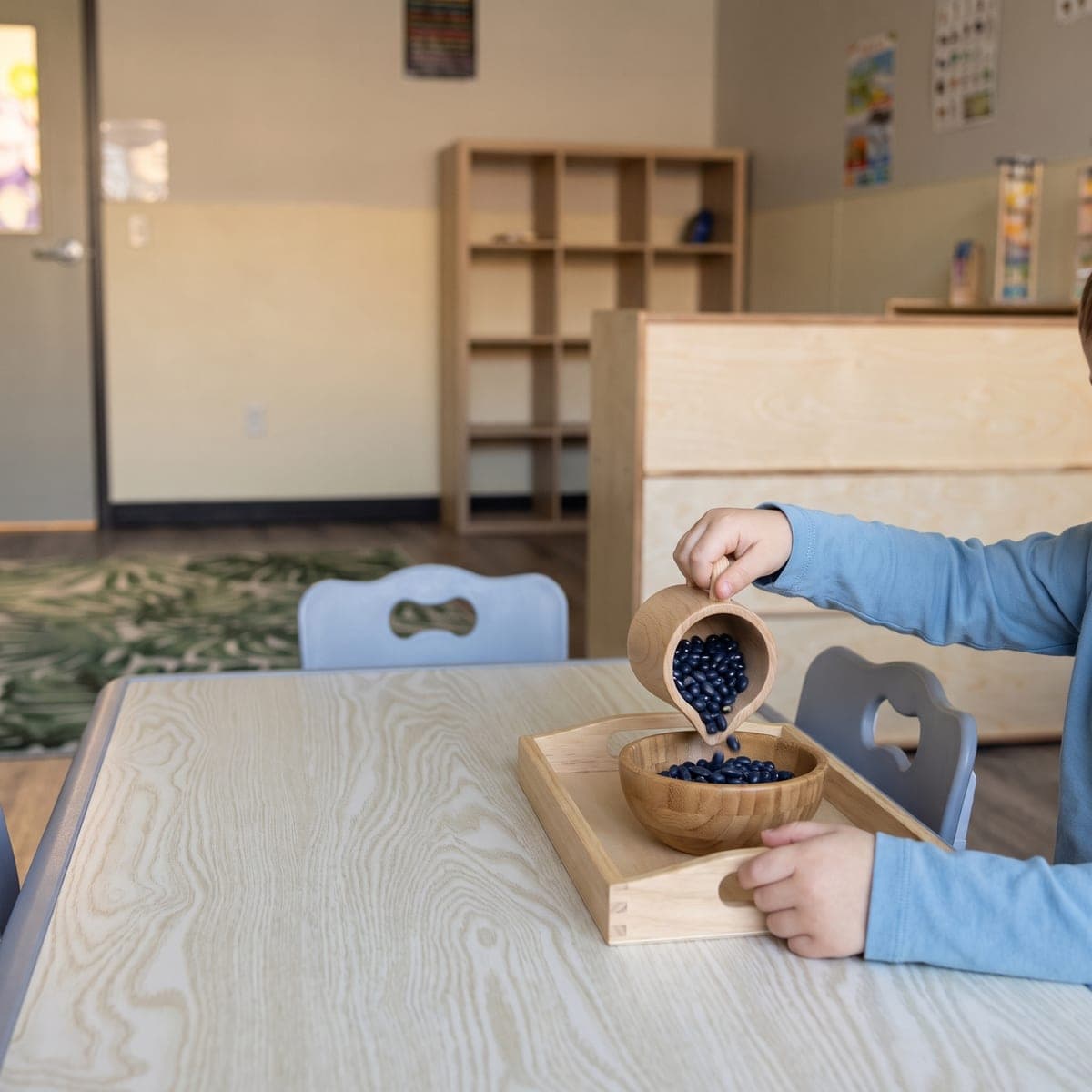 A child's hands pouring dry beans from a small pitcher into a bowl on a wooden tray