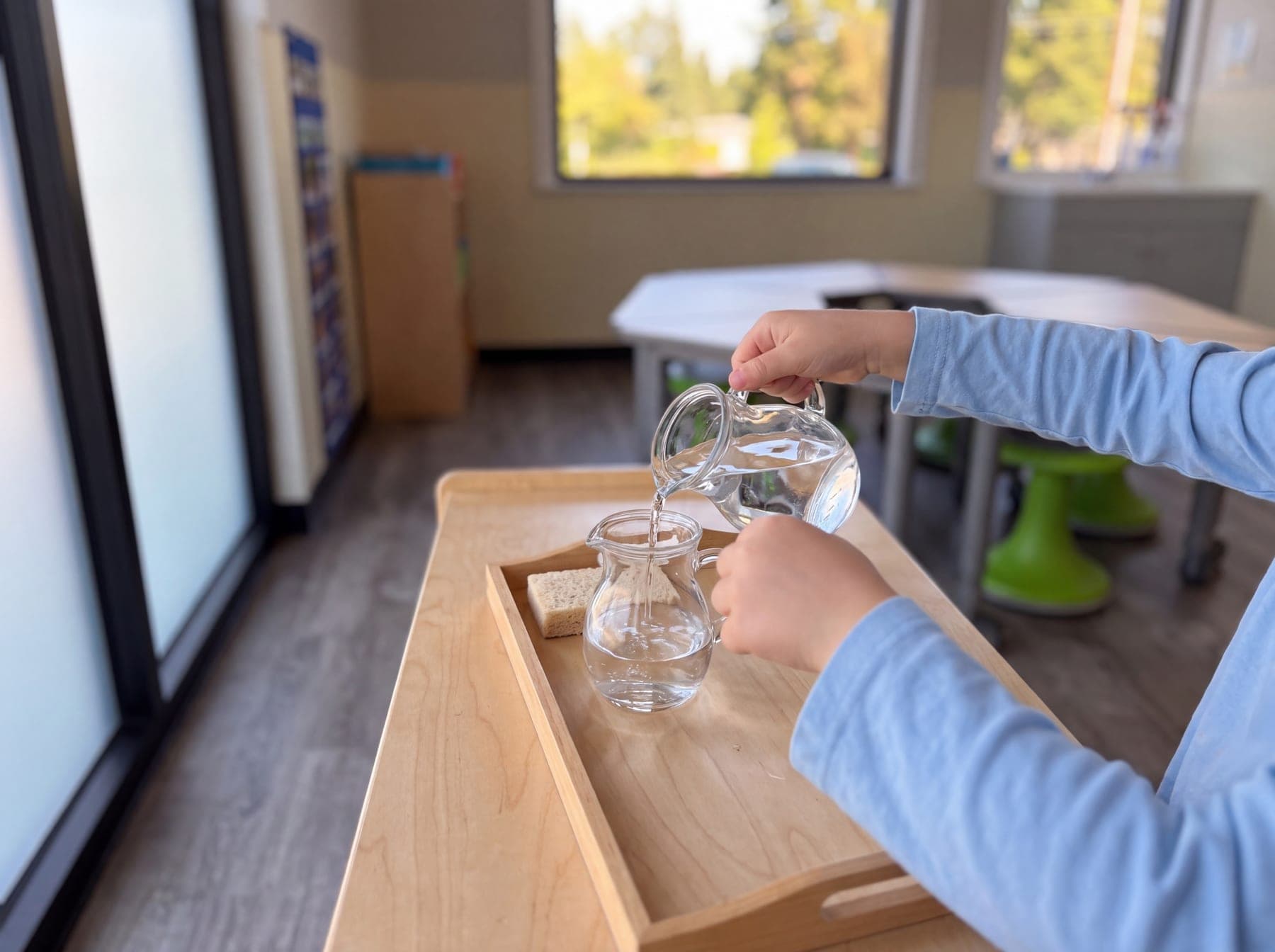A child's hands pouring water between two small glass pitchers on a wooden tray by the window